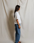 A woman stands in profile against a light beige fabric backdrop, wearing the Perfectwhitetee French Terry SS Loose Sweatshirt, blue jeans, and black sandals. Her straight dark hair is worn down.
