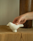 A hand gently places the Indaba Paper Mache Bird White accent decor on a wooden surface, with a tall wooden vase in the background.