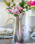 A Vintage Tall Tapered Pitcher by Faire, filled with pink and white flowers, sits on a set dining table featuring white plates, napkins, and additional floral centerpieces in the background.