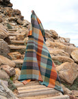 Outdoors on rocky steps, a person is mostly covered by the Vrou-Vrou Blanket from Faire, its green, orange, brown, and cream checks blending seamlessly into the rugged natural surroundings.