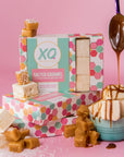 Boxes of Faire Salted Caramel Marshmallows are displayed on a pink background, surrounded by caramel pieces and a spoon drizzling sauce over allergy-friendly marshmallows stacked in a small bowl.
