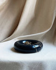 The Faire Gyrus Pocket Tray, a glossy black concrete piece with a cork base, holds rings and a gold necklace atop cream textured fabric with soft folds, offering both style and surface protection.