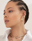 A woman with braided hair wears several gold earrings—including Faire’s Lisa Ear Cuff in 10K gold on her mid-ear cartilage—plus layered gold necklaces and a white collared shirt, glancing to the side against a neutral background.