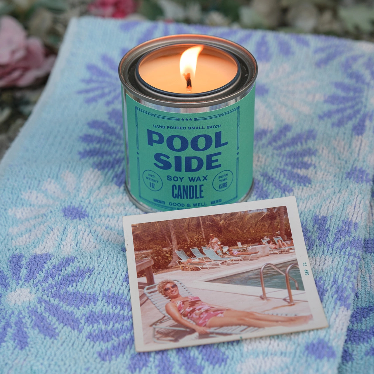 A lit G&W Soy Candle by Faire sits on a blue and purple patterned towel next to a vintage photo of a woman in sunglasses and a swimsuit, evoking nostalgic joy by the pool.