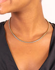 A woman wearing a black v-neck top and the Faire Turquoise Tennis Necklace, featuring small white beads and made from hypoallergenic 316L stainless steel, shown from chin to chest.