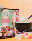 Boxes of Faire Vanilla Marshmallows are stacked with loose marshmallows, Madagascar vanilla beans, and a bowl of vanilla ice cream, all arranged on a bright yellow background.