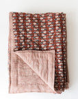 The Blossom Block Print Quilt by Faire features a brown background with white and blue floral prints on one side and thin red and white stripes on the reverse, shown with a turned-up corner to display both artisan-crafted patterns.