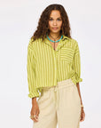A woman in the Kerri Rosenthal Tia Faded Stripe Shirt—lime green with a pocket—beige drawstring pants, turquoise necklaces, and rings stands against a plain white background.