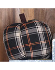 The Faire Pumpkin is a fabric ornament with black, beige, and orange plaid and a brown stem. Shown on a metal surface against wood, it makes the perfect farmhouse decor accent for autumn.