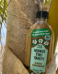 A bottle of Monoi Coco Coconut Oil by Faire leans against a palm tree trunk, wrapped in burlap, with lush green palms and sandy beach behind—showcasing the pure essence of Monoi Oil from Faire.