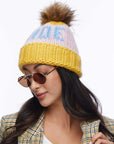 A woman in round sunglasses and a Faire Knit Ski Hat with a brown faux fur pom pom, paired with a plaid jacket, looks down slightly with a neutral expression against a white background.