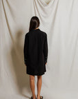 A woman with long hair stands facing away, wearing the Perfectwhitetee Felicia Poplin Dress and black sandals against draped white fabric—an ideal choice for effortless everyday style.