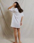 A woman with long, wavy brown hair faces away in the Felicia Poplin Dress by Perfectwhitetee—an oversized white cotton shirt dress paired with black flip-flops, set against a neutral backdrop. Ideal for effortless, everyday style.