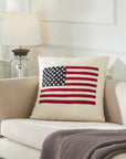 The Faire American Flag Wool Pillow, in cream felted wool and hand-embroidered, sits on a beige sofa beside a gray blanket, with a glass-based lamp visible on a table in the background.