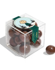The Faire Christmas Candy Cube is a clear box of chocolate-covered balls with a Charlie Brown Peanuts sticker on the lid—one ball outside. Ideal for sharing assorted candies or gifting as cheerful holiday sweets.