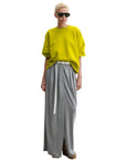 Wearing the Rachel Comey Fond Sweatshirt in bright yellow, a person pairs it with a gray maxi skirt with a front slit, white belt, brown shoes, and black sunglasses, standing against a white background.