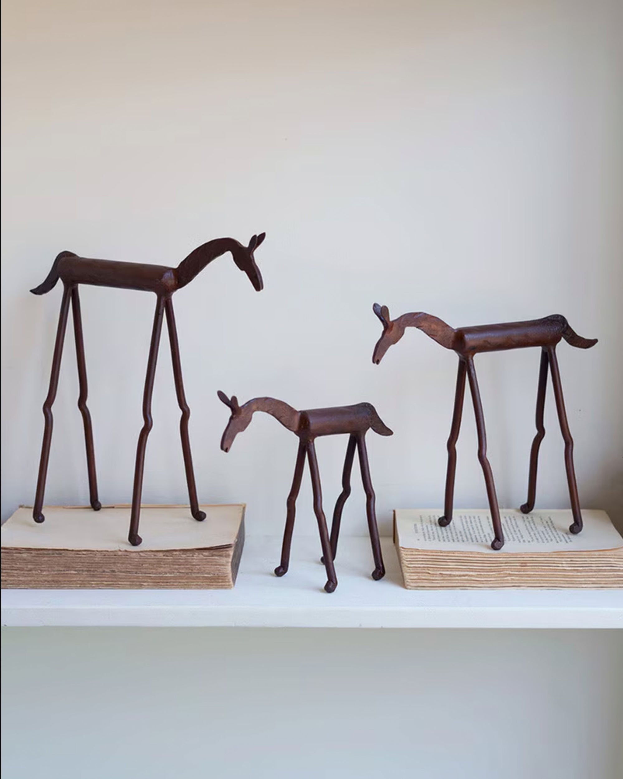 Three Kalalou, Inc Forged Iron Horse figurines with elongated legs are displayed on or beside old hardcover books on a shelf against a plain, light-colored background. These elegant pieces are handcrafted in India.