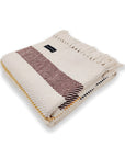 A folded cream-colored Faire Frontier Cotton Throw with brown and yellow stripes, fringed edges, and a black fabric label is shown on a white background.