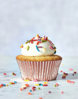 A Wild Rye Frosting plant-based vanilla cupcake with white vanilla frosting and colorful sprinkles sits in a shiny pink wrapper on a marble surface, with extra sprinkles scattered around.