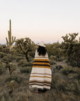 Faire Golden Horizon Handwoven Blanket from Bungalow Furniture draped on person outdoors among cacti at sunset.
