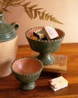 Two Green Terra Clay Compote bowls by Kalalou, Inc sit on a wooden surface—one holds decorative soap bars. A beige vase with ferns stands nearby, and a soap bar rests in front of the bowls.
