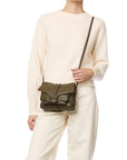 A person in off-white sweater and pants holds the Jerome Dreyfuss Harry Handbag, a dark olive green goatskin crossbody with a flap, buckle detail, and adjustable strap, standing against a plain white background.