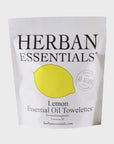 A white package labeled "Herban Essentials" contains Essential Oil Towelettes. The design features a yellow lemon illustration and mentions "Aromatherapeutic" with 20 towelettes included. The text also highlights "All Natural," "Clean Up, Calm Down," and hints of eucalyptus essential oil for added refreshment.
