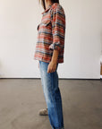 A person stands sideways in a room, wearing Hey Gang’s The Work Shirt in rust plaid with rolled sleeves, paired with blue jeans and gray shoes. The background has white walls and a smooth gray floor.