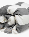 The Hedgehouse Throwbed features wide horizontal gray and white stripes, a small green tassel on one seam, and a removable cotton cover for easy care.
