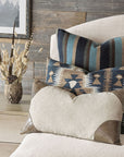 A beige chair displays three HIGGINS LEATHER CORNERS pillows by Eastern Accents with blue, brown, and Rasmus Khaki geometric and tribal designs. A rustic vase with dried grasses rests on a light wooden table nearby.