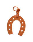 The Faire Bag Charm is a textured orange leather, horseshoe-shaped accessory with square cutouts and a matching hanging loop, adding a stylish, personal touch. Displayed on a white background.