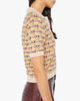 A woman is pictured in profile, wearing Mother’s The Hot Under the Collar Polo—a yellow short-sleeve knit top with pink and purple checkerboard accents—paired with high-waisted brown leather pants, her head gently inclined downward.