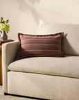 A cream sofa features the Loloi Rugs PCJ0023 Howie Wine Pillow 13x21, showcasing maroon and beige stripes. Next to it stands a wooden side table with an antique bronze finish and a flower vase against a light wall.