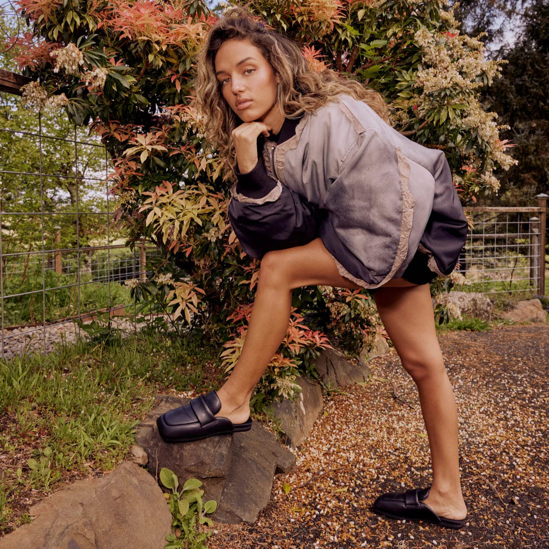 A woman with curly hair poses outdoors in an oversized gray jacket, black shorts, and Roam's Puffy Loafer mule loafers, resting one foot on a rock to highlight their memory foam insoles.