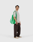 A man stands against a plain background, wearing a white T-shirt, dark brown pants, white sneakers, and carrying the large green and white striped Big Baggu recycled ripstop nylon shopping bag by Baggu over his shoulder.