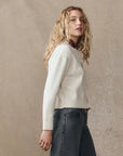 A woman with long, wavy blonde hair stands sideways in The Great Inc. Beaded Fringe Cardigan and high-waisted gray jeans against a plain beige background.
