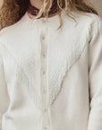 A person with wavy blonde hair wears The Great Inc. Beaded Fringe Cardigan, a cream knit with textured chevron patterns and beaded fringe, buttoned down the front, styled with a maroon bandana around their neck.