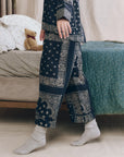 Someone wearing The Great Inc. Easy Sleep Pant in blue bandana patchwork print with gray socks stands by a bed featuring green and white bedding, a teddy bear resting on the pillows.