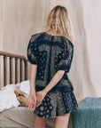 A woman with wavy blonde hair stands with her back to the camera, wearing The Sleep Short by The Great Inc.—a navy blue, bandana patchwork print short—in a bedroom with floral bedding and a teddy bear.