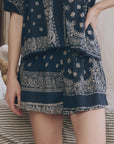 A person models The Great Inc.’s The Sleep Short—a matching navy blue set with a white paisley floral bandana patchwork print—posing with one hand on their hip against a neutral background.