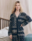 A woman with wavy blonde hair stands indoors by a wooden bed, wearing The Pajama Shirt by The Great Inc.—a vintage-inspired, navy blue and white patterned women's sleepwear top—with one hand on her hip and a neutral expression.