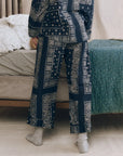 A person wearing The Great Inc. Easy Sleep Pant in high waist navy blue cotton with a white bandana patchwork print stands by a bed with striped and floral bedding, light gray socks completing the cozy look.