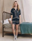 A woman with wavy blonde hair smiles in front of a vintage metal bed, wearing The Great Inc.'s The Sleep Short in navy Bandana Patchwork Print with an elastic waist and gray socks. The bed has blue and white bedding and a teddy bear on the pillows.