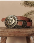 The Leather Concho Belt by The Great Inc., featuring tooled black leather, embossed patterns, and two large ornate brass buckles, is displayed on a rustic wooden stool against a beige background.