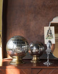 Two Blue Ocean Traders Wunderkammer Orbs on brass stands sit on wood beside a mirror held by a silver hand, which displays a vintage portrait card. A brown metal backdrop frames this distinctive vintage scene.