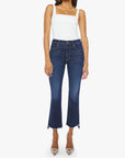 A woman stands against a plain white background, wearing a white sleeveless square-neck top, Mother’s The Insider Crop Step Fray dark blue high-waisted bootcut jeans with a frayed hem, and pointed clear heels.