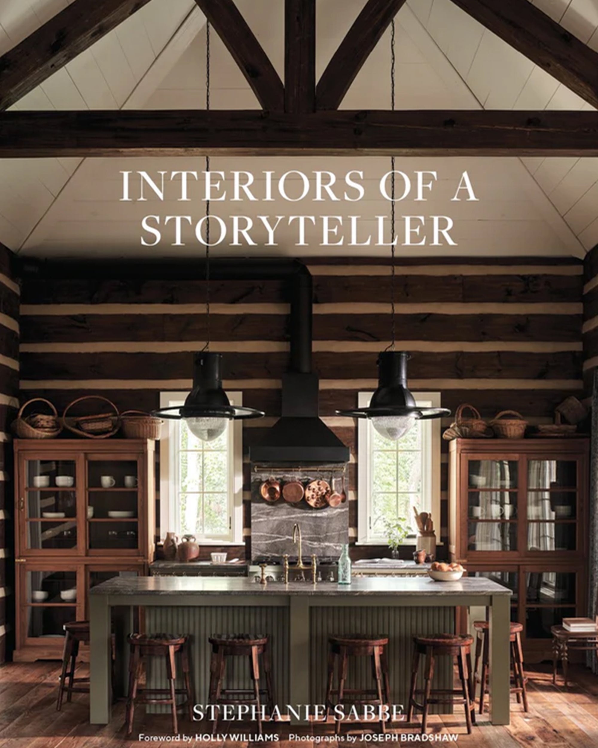 A rustic kitchen with wood beams, a large island and stools, glass cabinets, pendant lights, and sunlit windows features in "Interiors of a Storyteller" by Gibbs Smith Publisher.