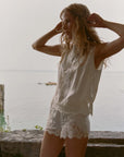 A woman wearing the DOEN Iona Short and a sleeveless floral lace white top stands outdoors by stonework with water and a hazy sky behind her, adjusting her hair and gazing to the side.