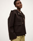 A person with short hair wears the loose-fitting, dark BA&SH Jacket Kove with large pockets and decorative buttons, paired with light green pants, standing against a plain light background.