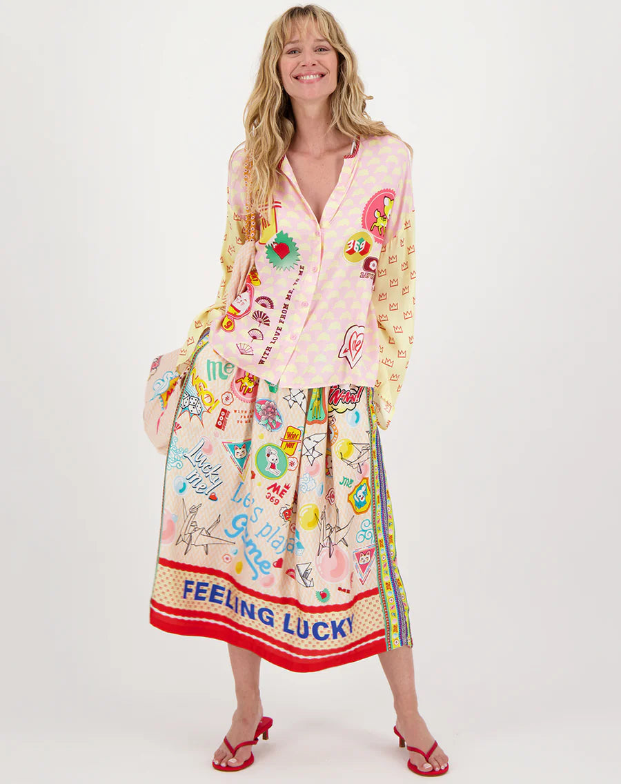 A smiling woman wears a cheerful ensemble featuring hearts, cherries, and flowers. She dons the Me369 Vanessa Midi Skirt with "FEELING LUCKY" at the hem, complete with a matching shirt and red sandals.